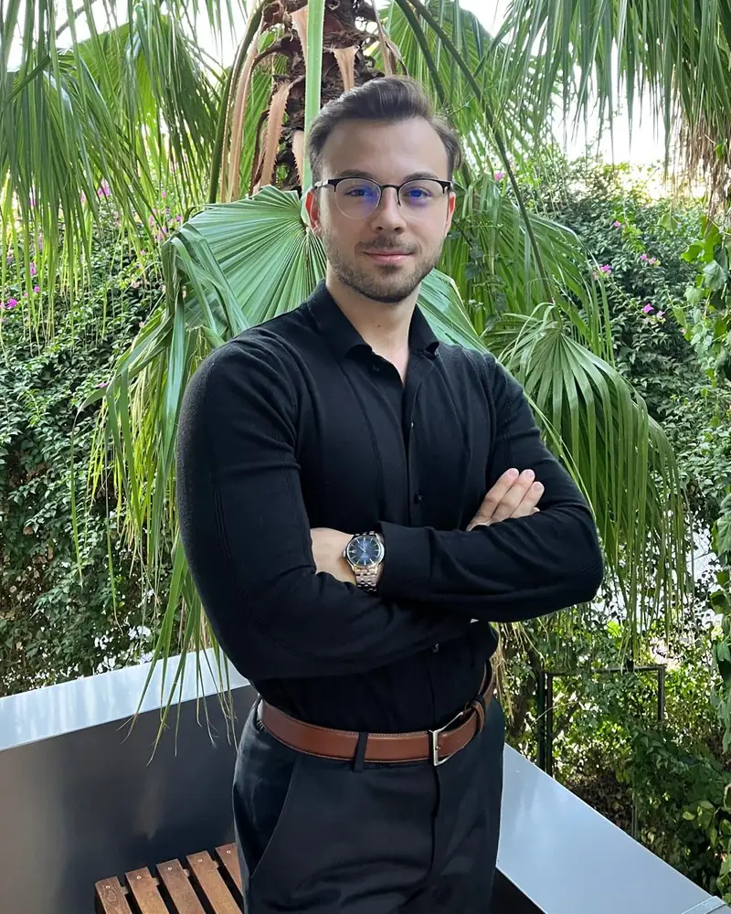 Diyetisyen Arman Ünsal, yeşil bitkilerin önünde kollarını kavuşturmuş halde